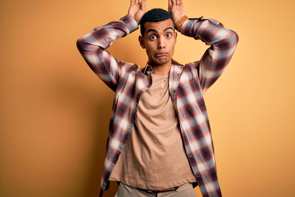 Jonge knappe Afro-Amerikaanse man draagt casual shirt staan over gele achtergrond Doen konijnenoren gebaar met handen palmen kijken cynisch en sceptisch. Paashaas concept. - Foto, afbeelding