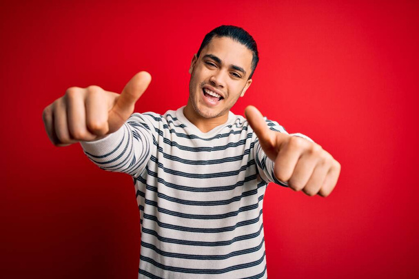 Jeune homme brésilien portant casual t-shirt rayé debout sur fond rouge isolé approuvant faire geste positif avec la main, pouces levés souriant et heureux pour le succès. Le geste du gagnant. - Photo, image