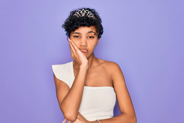 Nuori kaunis afrikkalainen afro nainen yllään tiara kruunu yli violetti tausta ajattelu näyttää väsynyt ja kyllästynyt masennus ongelmia ristissä kädet
. - Valokuva, kuva