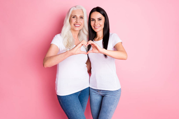 Photo of adorable aged mother young daughter hugging showing fingers heart figure symbolizing cardiac health wear white t-shirts jeans isolated pastel pink color background - Foto, Imagem