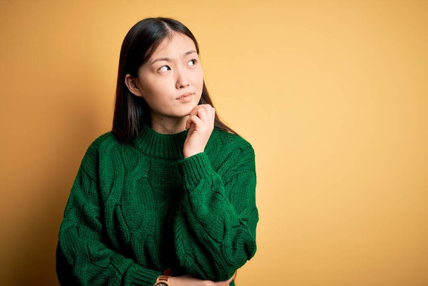 Jeune belle femme asiatique portant un pull d'hiver vert sur fond jaune isolé avec la main sur le menton penser à la question, expression coûteuse. Souriant avec un visage réfléchi. Concept de doute
. - Photo, image