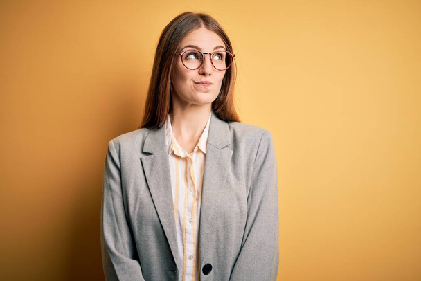 Young beautiful redhead woman wearing jacket and glasses over isolated yellow background smiling looking to the side and staring away thinking. - Fotografie, Obrázek