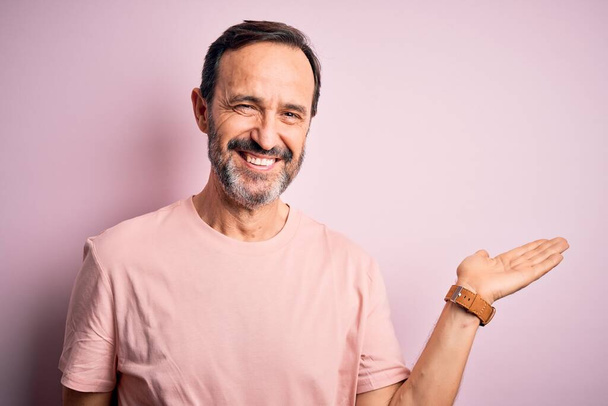 Middle age hoary man wearing casual t-shirt standing over isolated pink background smiling cheerful presenting and pointing with palm of hand looking at the camera. - Photo, Image