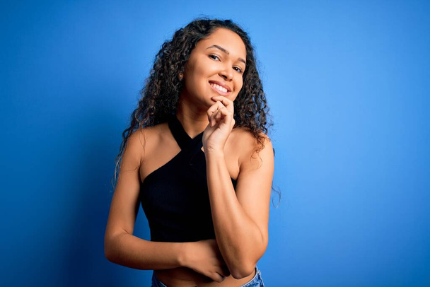 Young beautiful woman with curly hair wearing casual t-shirt standing over blue background looking confident at the camera with smile with crossed arms and hand raised on chin. Thinking positive. - Photo, Image