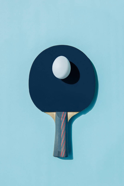 A white egg lies on a blue ping pong racket. A shadow falls from the egg. The handle of the racket has red stripes.  - Foto, Imagem