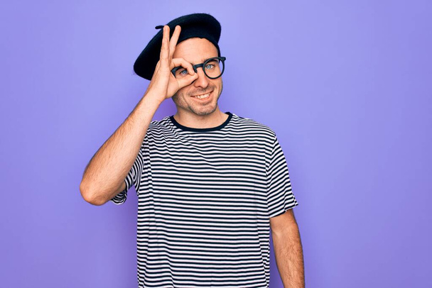Handsome man with blue eyes wearing striped t-shirt and french beret over purple background doing ok gesture with hand smiling, eye looking through fingers with happy face. - 写真・画像