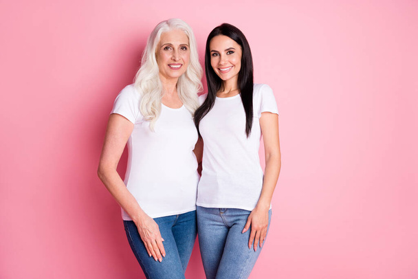 Photo of cheerful pretty aged mother young daughter ladies generation hugging spend time together wear white t-shirts jeans isolated pastel pink color background - Photo, image