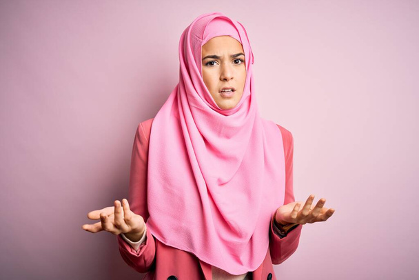 İslami tesettür giyen genç ve güzel bir kız pembe arka planda duruyor. Hiçbir şeyden habersiz ve kolları açık, hiçbir fikri yok.. - Fotoğraf, Görsel