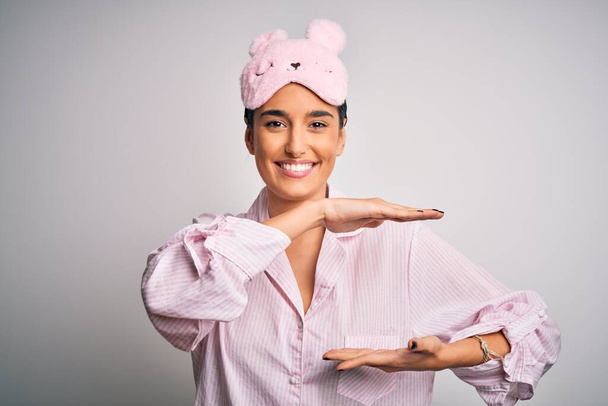 Young beautiful brunette woman wearing pajama and sleep mask over white background gesturing with hands showing big and large size sign, measure symbol. Smiling looking at the camera. Measuring concept. - Fotó, kép