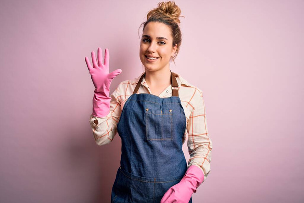Joven hermosa mujer rubia limpiadora haciendo tareas domésticas con arpon y guantes mostrando y señalando con los dedos número cinco mientras sonríe confiado y feliz
. - Foto, Imagen