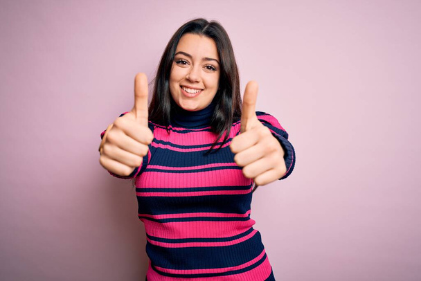 ピンクの隔離された背景の上に縞模様のシャツを身に着けている若いブルネットのエレガントな女性は、手で肯定的なジェスチャーを行うことを承認し、親指を笑顔と成功のために幸せ。勝者のジェスチャー. - 写真・画像