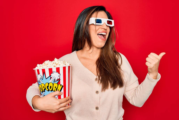 Nuori latino nainen yllään 3D-elokuva lasit ja syö popcornia yli punainen tausta osoittaa ja osoittaa peukalolla jopa puolelle onnellinen kasvot hymyillen
 - Valokuva, kuva
