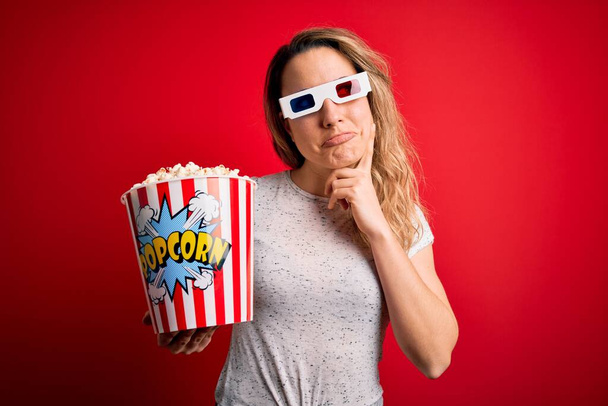 jong mooi blond vrouw kijken film met behulp van 3D bril eten popcorn snack serieus gezicht denken over vraag, zeer verward idee - Foto, afbeelding