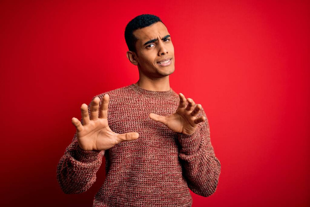 Young handsome african american man wearing casual sweater standing over red background disgusted expression, displeased and fearful doing disgust face because aversion reaction. With hands raised - Valokuva, kuva