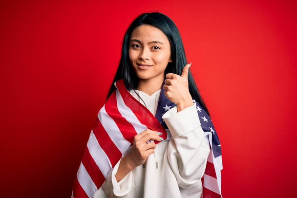 Joven hermosa mujer china con bandera de los Estados Unidos sobre fondo rojo aislado feliz con una gran sonrisa haciendo signo ok, pulgar hacia arriba con los dedos, señal excelente
 - Foto, imagen