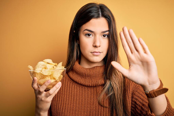 Young beautiful girl holding bowl with chips potatoes standing over yellow background with open hand doing stop sign with serious and confident expression, defense gesture - Foto, Bild