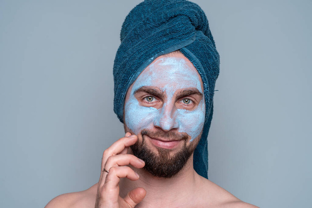 Бородатий чоловік з блакитним рушником на голові і косметичною маскою на обличчі. Косметологічні процедури для чоловіків. Догляд за шкірою для чоловіків
. - Фото, зображення