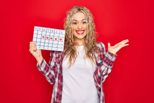 Joven hermosa mujer rubia sosteniendo el calendario del período que controla el ciclo menstrual celebrando la victoria con sonrisa feliz y expresión ganadora con las manos levantadas
 - Foto, imagen
