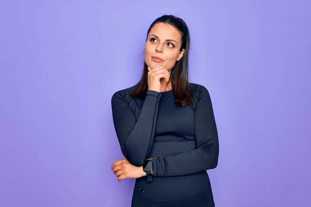 Young beautiful brunette sporty woman wearing casual sportswear over purple background with hand on chin thinking about question, pensive expression. Smiling with thoughtful face. Doubt concept. - Zdjęcie, obraz