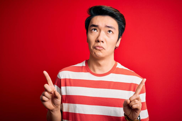 Fiatal jóképű kínai férfi, alkalmi csíkos pólót visel, piros háttér felett áll, szomorúan és feldúltan néz, jelezve az irányt az ujjaival, boldogtalan és depressziós. - Fotó, kép