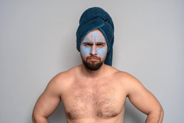 頭に青いタオルを被った髭の男と顔に化粧品マスク。男性用化粧品の手順.男性用スキンケア. - 写真・画像