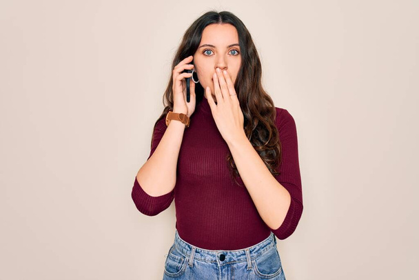 Mladá krásná žena vede rozhovor na smartphonu přes bílé pozadí kryt úst s rukou šokován hanbou za omyl, výraz strachu, strach v tichu, tajný koncept - Fotografie, Obrázek
