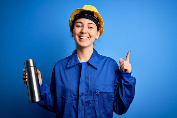 Nuori kaunis brunette työntekijä nainen yllään turvallisuus kypärä tilalla termo vedellä yllättynyt idea tai kysymys osoittaa sormella onnellinen kasvot, numero yksi
 - Valokuva, kuva