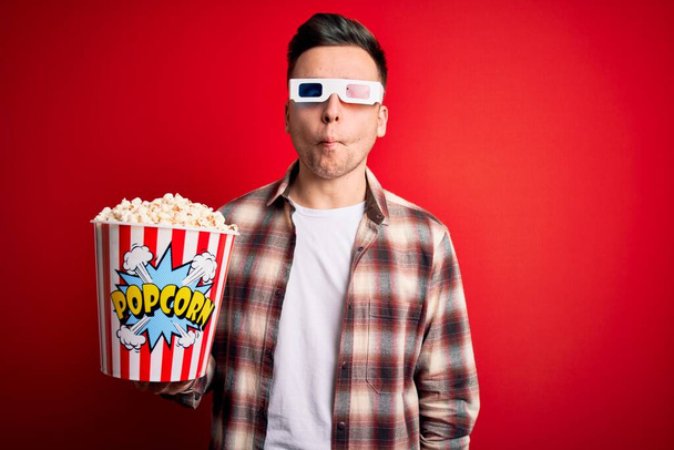 Young handsome caucasian man wearing 3d movie glasses and eating popcorn making fish face with lips, crazy and comical gesture. Funny expression. - Photo, Image