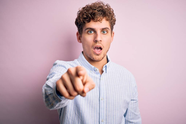 Young blond handsome man with curly hair wearing striped shirt over white background pointing displeased and frustrated to the camera, angry and furious with you - Photo, Image