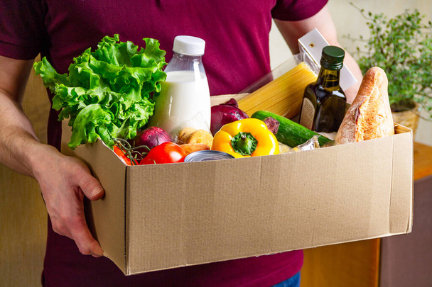 Volunteer holding food in a donation cardboard box with various food. Open cardboard box with oil, vegetables, milk, canned food, cereals and pasta. Food delivery concept with copy space - 写真・画像