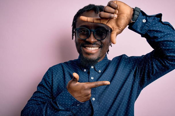 Jonge knappe Afro-Amerikaanse man draagt casual shirt en bril over roze achtergrond glimlachende maken frame met handen en vingers met gelukkig gezicht. Creativiteit en fotografie concept. - Foto, afbeelding