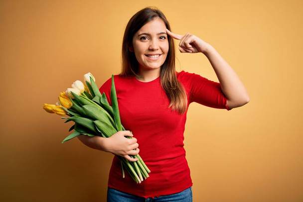 Mulher loira jovem segurando buquê romântico de flores de tulipas sobre fundo amarelo Sorrindo apontando para a cabeça com um dedo, ótima ideia ou pensamento, boa memória
 - Foto, Imagem