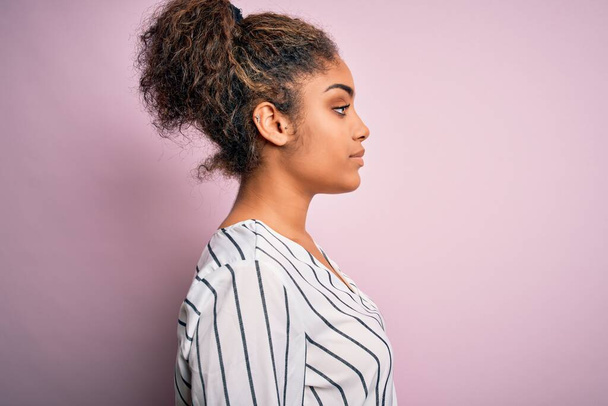 Jeune belle fille afro-américaine portant t-shirt rayé debout sur fond rose regardant vers le côté, pose de profil relax avec visage naturel avec sourire confiant
. - Photo, image