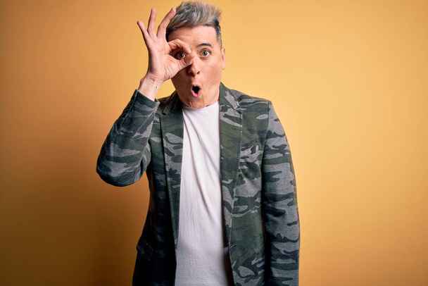 Young handsome modern man wearing business jacket standing over yellow isolated background doing ok gesture shocked with surprised face, eye looking through fingers. Unbelieving expression. - Фото, изображение
