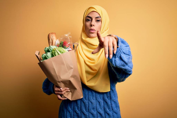 Fiatal gyönyörű barna arab nő visel iszlám hidzsáb tartó papírzacskó élelmiszer mutató ujjal a kamera és az Ön számára, kézjel, pozitív és magabiztos gesztus az első - Fotó, kép