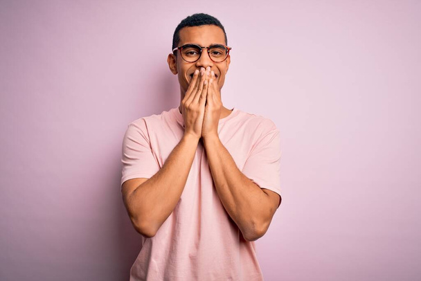 Pohledný africký Američan nosí ležérní tričko a brýle přes růžové pozadí se smíchem a rozpačité hihňání zakrývající ústa s rukama, drby a skandální koncept - Fotografie, Obrázek