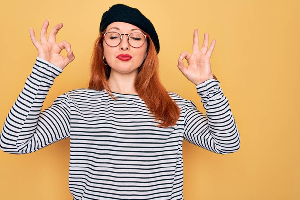 Beautiful redhead woman wearing striped t-shirt and french beret over yellow background relax and smiling with eyes closed doing meditation gesture with fingers. Yoga concept. - Фото, зображення