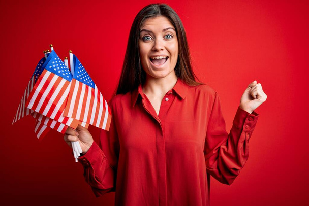 Νεαρή όμορφη πατριώτισσα με μπλε μάτια που κρατά σημαίες των Ηνωμένων Πολιτειών πάνω από κόκκινο φόντο ουρλιάζοντας περήφανη και γιορτάζοντας τη νίκη και την επιτυχία πολύ ενθουσιασμένος, ζητωκραυγάζοντας συγκίνηση - Φωτογραφία, εικόνα