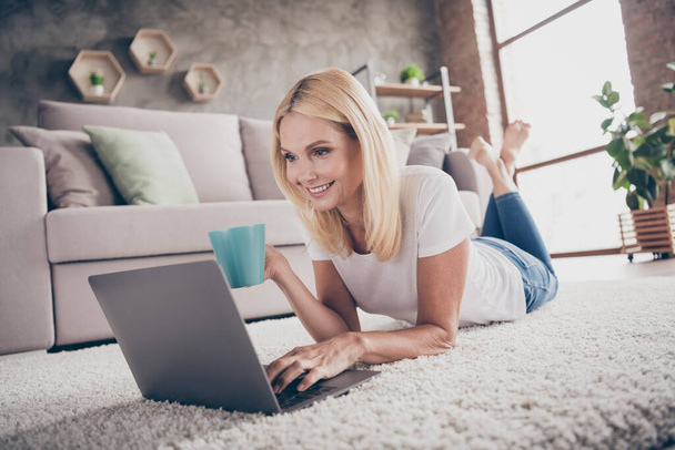 Close-up portrait of her she nice attractive charming cheerful focused mature lady lying on carpet drinking tea using laptop browsing in modern industrial loft interior style living-room apartment - Foto, Bild