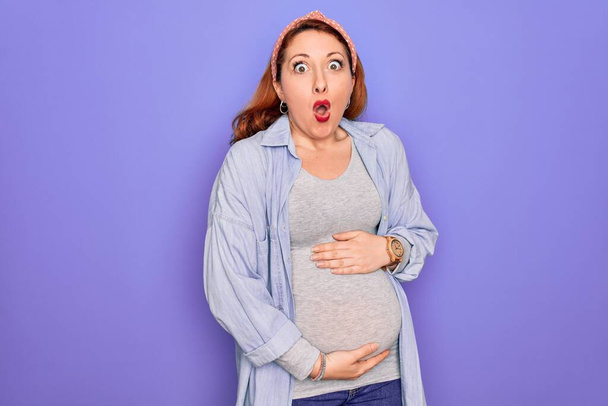 Νεαρή όμορφη κοκκινομάλλα έγκυος γυναίκα αναμένουν μωρό πάνω από απομονωμένο μωβ φόντο φοβισμένος και σοκαρισμένος από την έκφραση έκπληξη, το φόβο και ενθουσιασμένος πρόσωπο. - Φωτογραφία, εικόνα