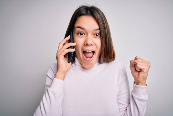 Beautiful brunette girl having conversation talking on the smartphone over white background screaming proud and celebrating victory and success very excited, cheering emotion - Zdjęcie, obraz