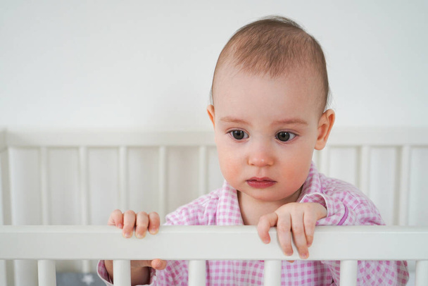 A small child in pink pajamas is standing in the crib. The kid woke up and waits for his parents to pull him out of the crib. Baby starts to cry - Photo, Image