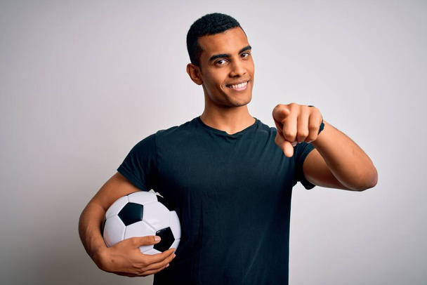 Przystojny Afroamerykanin gra w piłkę nożną trzymając piłkę na białym tle wskazując na ciebie i kamerę palcami, uśmiechając się pozytywnie i radośnie - Zdjęcie, obraz