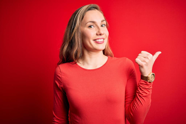 Young beautiful blonde woman wearing casual t-shirt standing over isolated red background smiling with happy face looking and pointing to the side with thumb up. - Фото, зображення