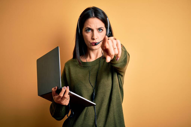 Молода жінка-оператор з блакитними очима працює на колл-центрі за допомогою комп'ютерного ноутбука і гарнітури, вказуючи пальцем на камеру і до вас, знак руки, позитивний і впевнений жест спереду
 - Фото, зображення