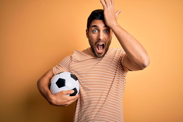 Przystojny gracz z brodą grający w piłkę nożną trzymający piłkę nożną na żółtym tle zaskoczony ręką na głowie przez pomyłkę, pamiętaj o błędzie. Zapomniałem, złe pojęcie pamięci. - Zdjęcie, obraz