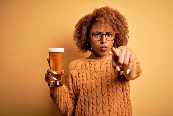 Jovem mulher afro afro afro afro bonita afro com cabelo encaracolado beber copo de cerveja apontando com o dedo para a câmera e para você, sinal de mão, gesto positivo e confiante da frente
 - Foto, Imagem