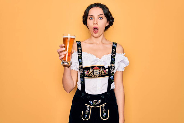 Junge schöne deutsche Frau mit blauen Augen im Oktoberfestkleid, die ein Glas Bier trinkt, erschrocken mit einem Überraschungsgesicht, ängstlich und aufgeregt mit Angstausdruck - Foto, Bild