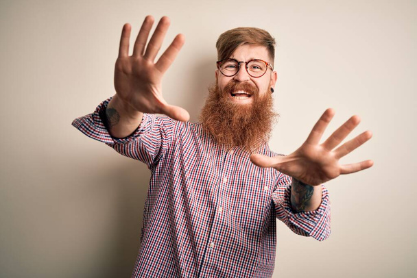 Przystojny irlandzki rudy biznesmen z brodą w okularach na odosobnionym tle, przerażony strachem, zatrzymuje gesty rękami, wrzeszcząc w szoku. Koncepcja paniki. - Zdjęcie, obraz