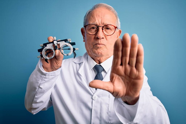 Senior γκρίζα μαλλιά οπτικός γιατρός άνθρωπος κρατώντας οπτομέτρη γυαλιά πάνω από το μπλε φόντο με ανοικτό χέρι κάνει σήμα στοπ με σοβαρή και σίγουρη έκφραση, χειρονομία άμυνας - Φωτογραφία, εικόνα